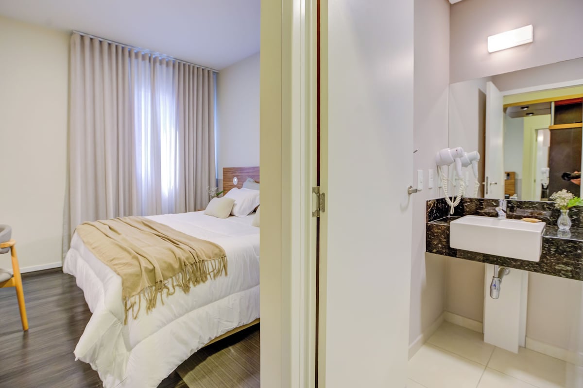 Une chambre d’hôtel avec un lit, une table de chevet, une chaise et une salle de bain équipée d’un lavabo et d’un miroir.