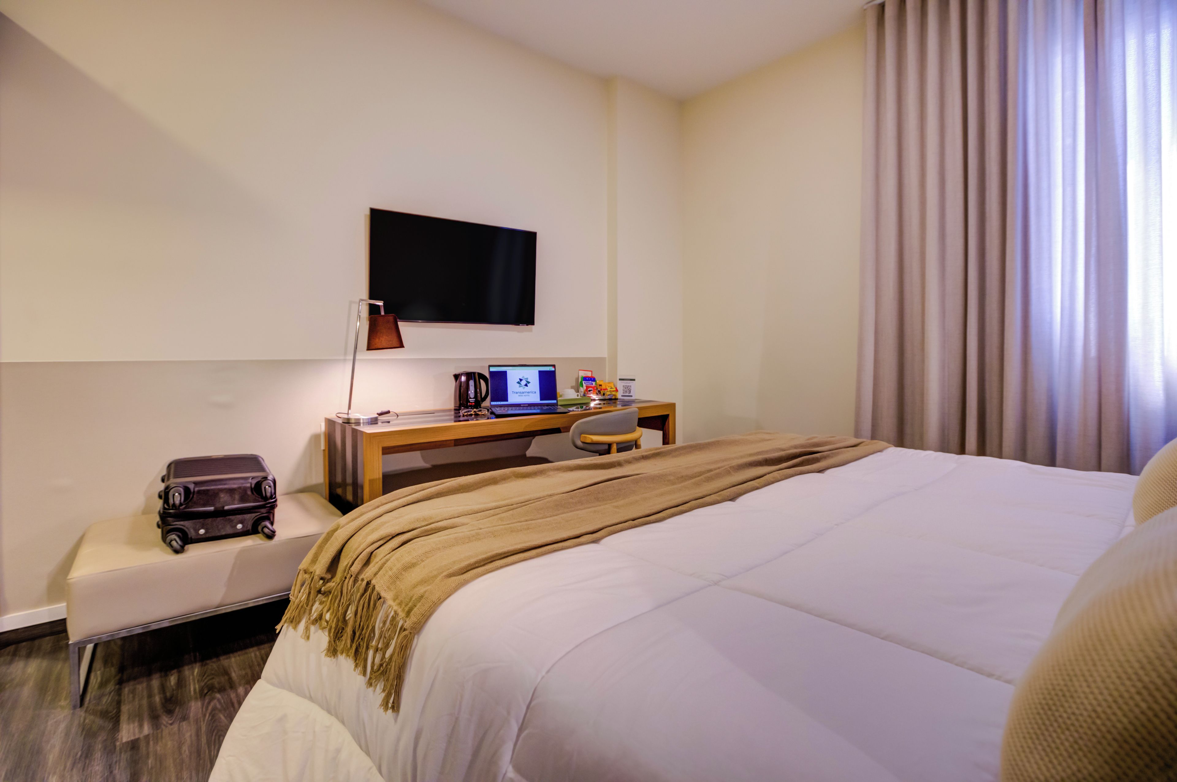 A hotel room with a bed, a desk with a laptop, a television, and a suitcase.