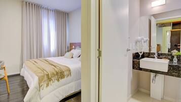 Quarto de hotel com cama, mesa de cabeceira, poltrona e banheiro com pia e espelho.