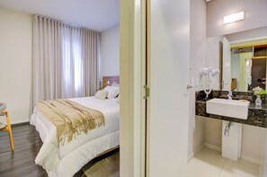 Een hotelkamer met een bed, nachtkastje, stoel en een badkamer met wastafel en spiegel.