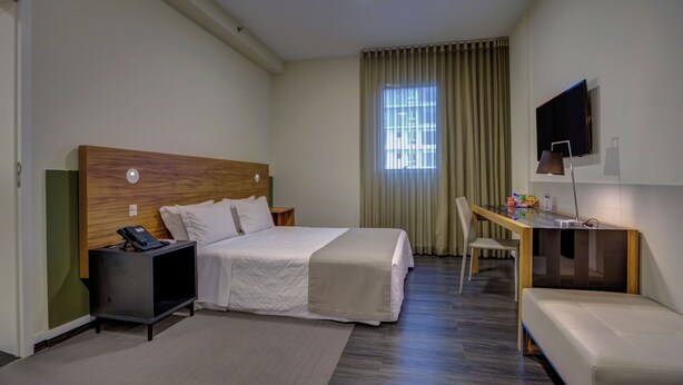 Une chambre d’hôtel comprenant un lit, une table de chevet avec un téléphone, un bureau avec une chaise, une télévision et une fenêtre avec des rideaux.