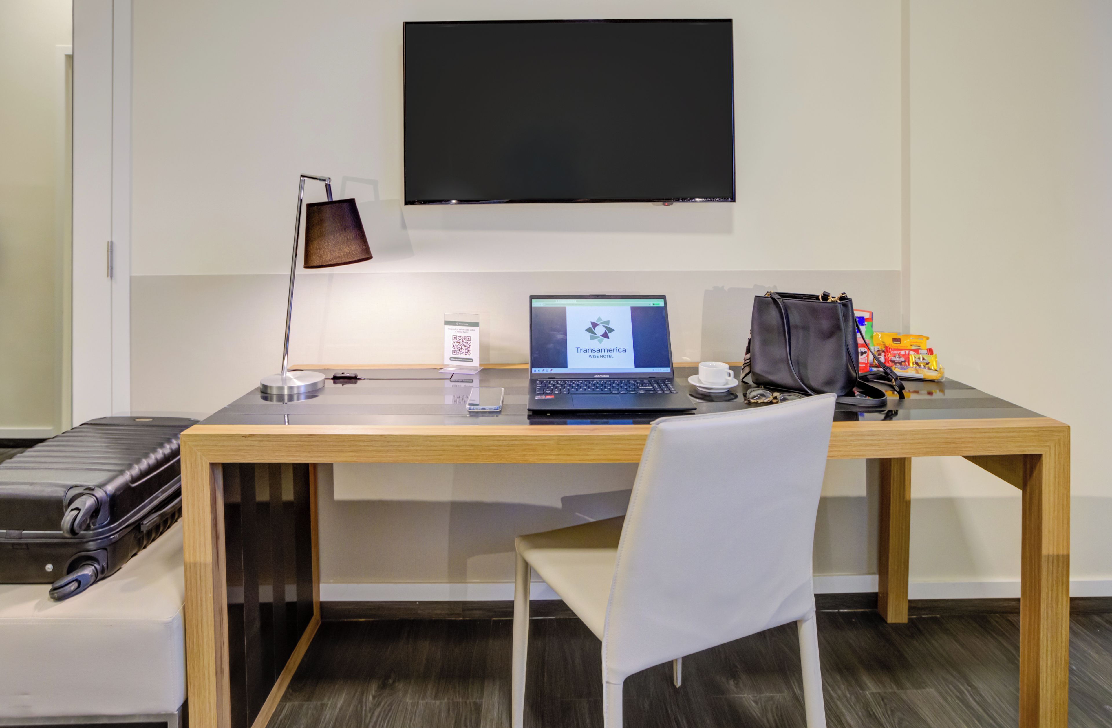 Habitación de hotel con escritorio de madera, una computadora portátil, una bolsa negra, una silla blanca y una maleta negra.