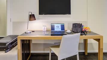 Habitación de hotel con escritorio de madera, una computadora portátil, una bolsa negra, una silla blanca y una maleta negra.