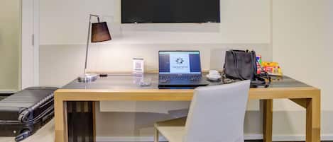 Habitación de hotel con escritorio de madera, una computadora portátil, una bolsa negra, una silla blanca y una maleta negra.