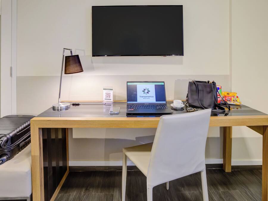 Une chambre d’hôtel avec un bureau en bois, un ordinateur portable, un sac noir, une chaise blanche et une valise noire.