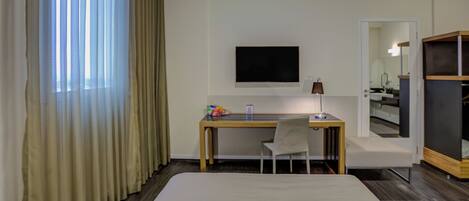 Una habitación de hotel con una cama, un escritorio con silla, un televisor y un espejo.