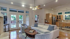 A spacious living room with a large sofa, a coffee table, and a ceiling fan. There is a kitchen area with wooden cabinets and a refrigerator. - 5BR Retreat in Bouldin Creek | Walk to SoCo (Austin)