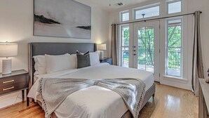 A bedroom with a large bed, a ceiling fan, a nightstand, a lamp, and a window with a view of greenery. - 5BR Retreat in Bouldin Creek | Walk to SoCo (Austin)