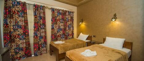 Habitación de hotel con dos camas individuales, una mesita de noche, una lámpara y cortinas estampadas.