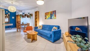 A living room with a blue sofa, wooden coffee table, and orange armchair. There is a TV, a bookshelf, and a painting on the wall. - Charming 2-Bedroom Apartment with Balconies MGAJ1-1 (Msida)