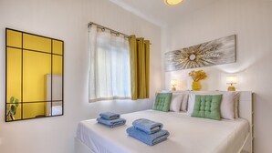A neatly made bed with folded towels, a large mirror, a window with curtains, and a wall-mounted artwork. - Charming 2-Bedroom Apartment with Balconies MGAJ1-1 (Msida)