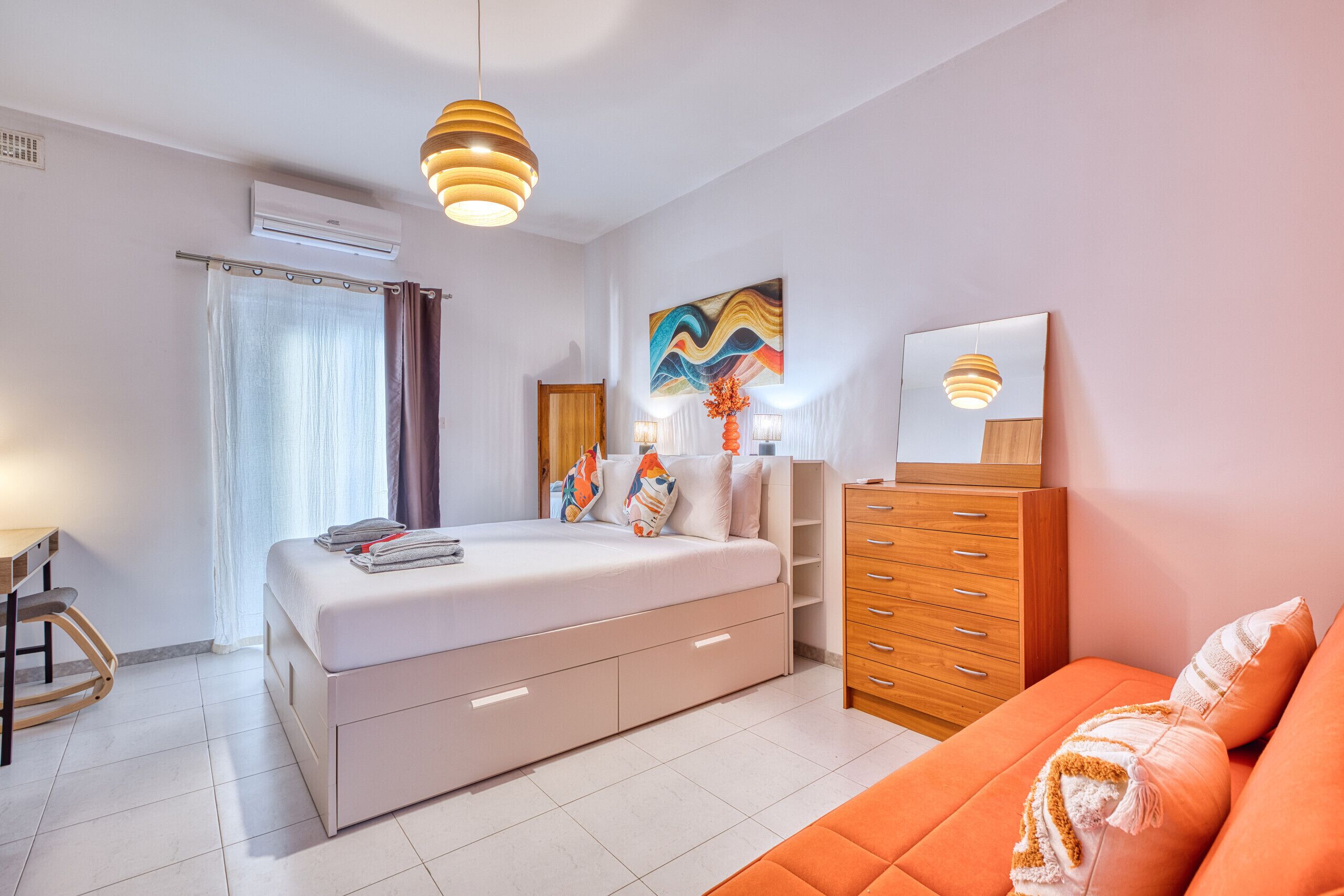 Een moderne slaapkamer met een groot bed, een houten dressoir en een oranje bank.