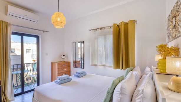 A hotel room with a bed, a dresser, a mirror, a window with curtains, and a balcony with a table and chairs. - Charming 2-Bedroom Apartment with Balconies MGAJ1-1 (Msida)