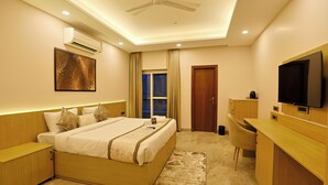 A hotel room with a bed, bedside table, desk, TV, air conditioning unit, and a window with curtains. - Millora Cyber Suites (Gurugram)