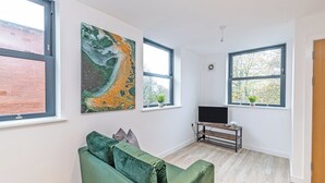 Een moderne woonkamer met een groene bank, kunstwerken aan de muren, een televisie op een houten dressoir en grote ramen met uitzicht op bomen.