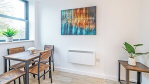 Een moderne eethoek met een houten tafel en stoelen, een groot abstract schilderij aan de muur, een radiator eronder en een raam met een plant op de vensterbank.