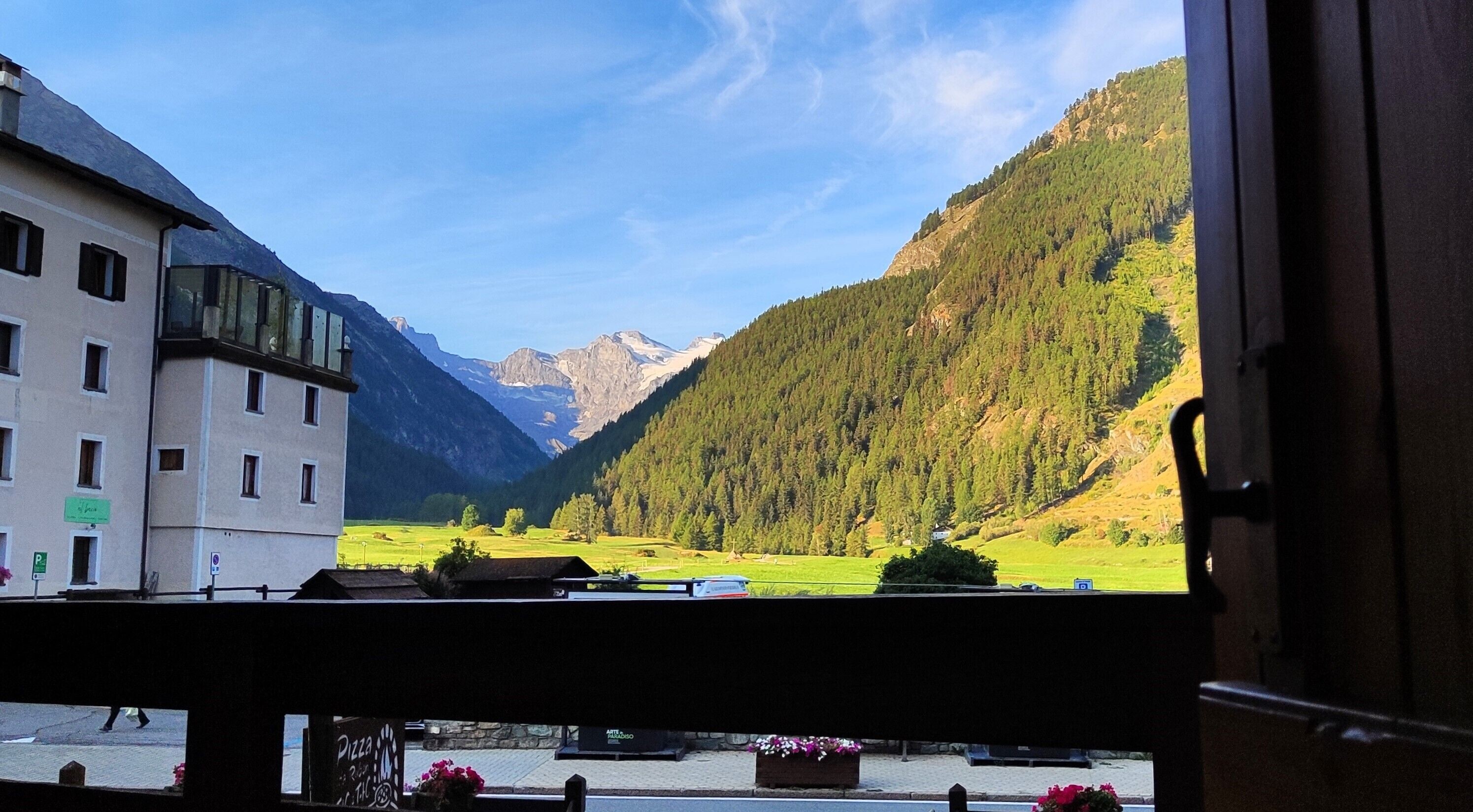 Udsigt fra en balkon over et dal landskab med bjerge, en bygning med et skilt der angiver en parkeringsplads, og en blomsterkasse med lyserøde blomster.