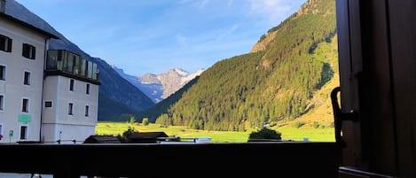 A view from a balcony overlooking a valley with mountains, a building with a sign indicating a parking area, and a flower box with pink flowers.