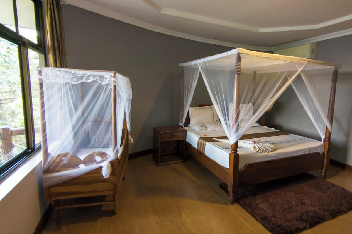 A room with two wooden four-poster beds with white bedding and mosquito netting. There is a wooden nightstand and a small wooden table beside each bed. The room has wooden flooring and a brown rug on the floor.