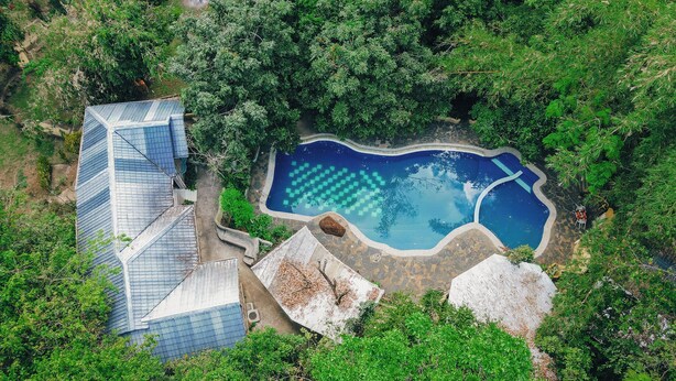 Vista aérea de un complejo turístico con piscina rodeada de abundante vegetación y un edificio moderno con techo inclinado.