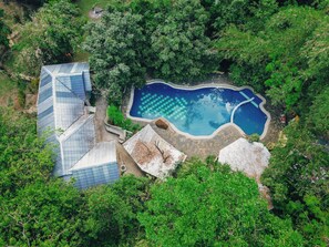 Vue aérienne d’un complexe hôtelier doté d’une piscine entourée d’une végétation luxuriante et d’un bâtiment moderne à toit en pente.