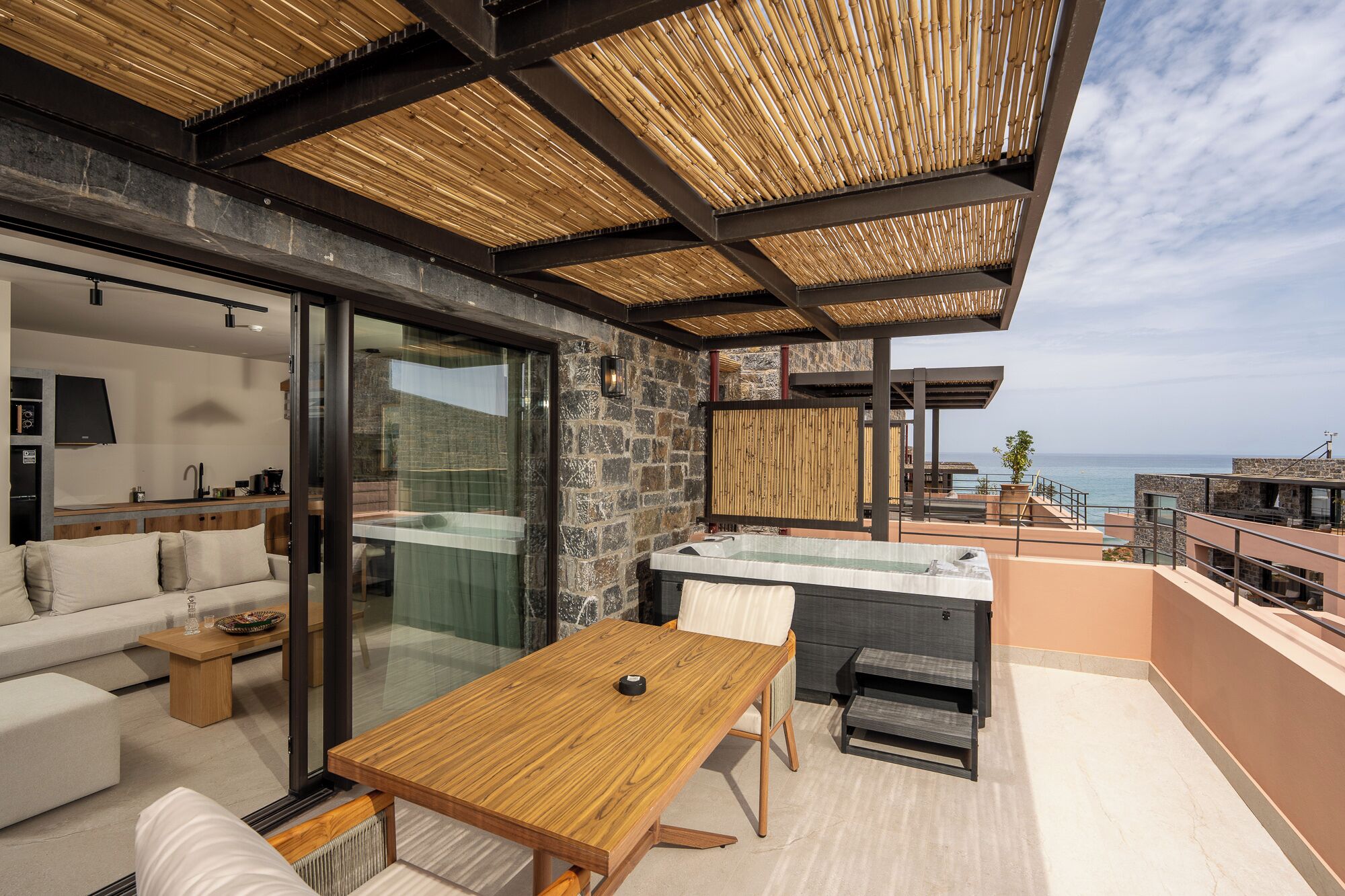 A modern outdoor living space with a wooden table, a sofa, a hot tub, and a stone wall.