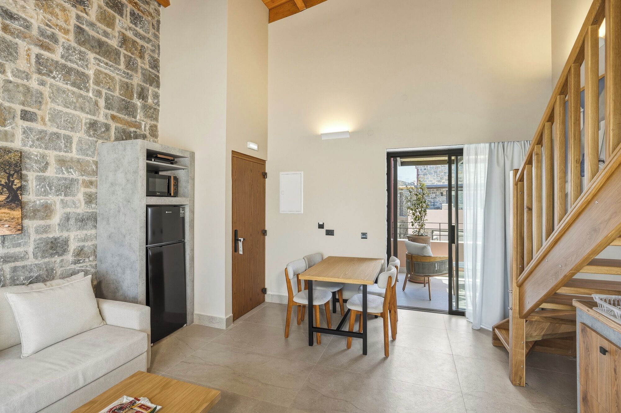 A living room with a stone wall, a wooden staircase, a dining area with a table and chairs, and a kitchen with a refrigerator and microwave.