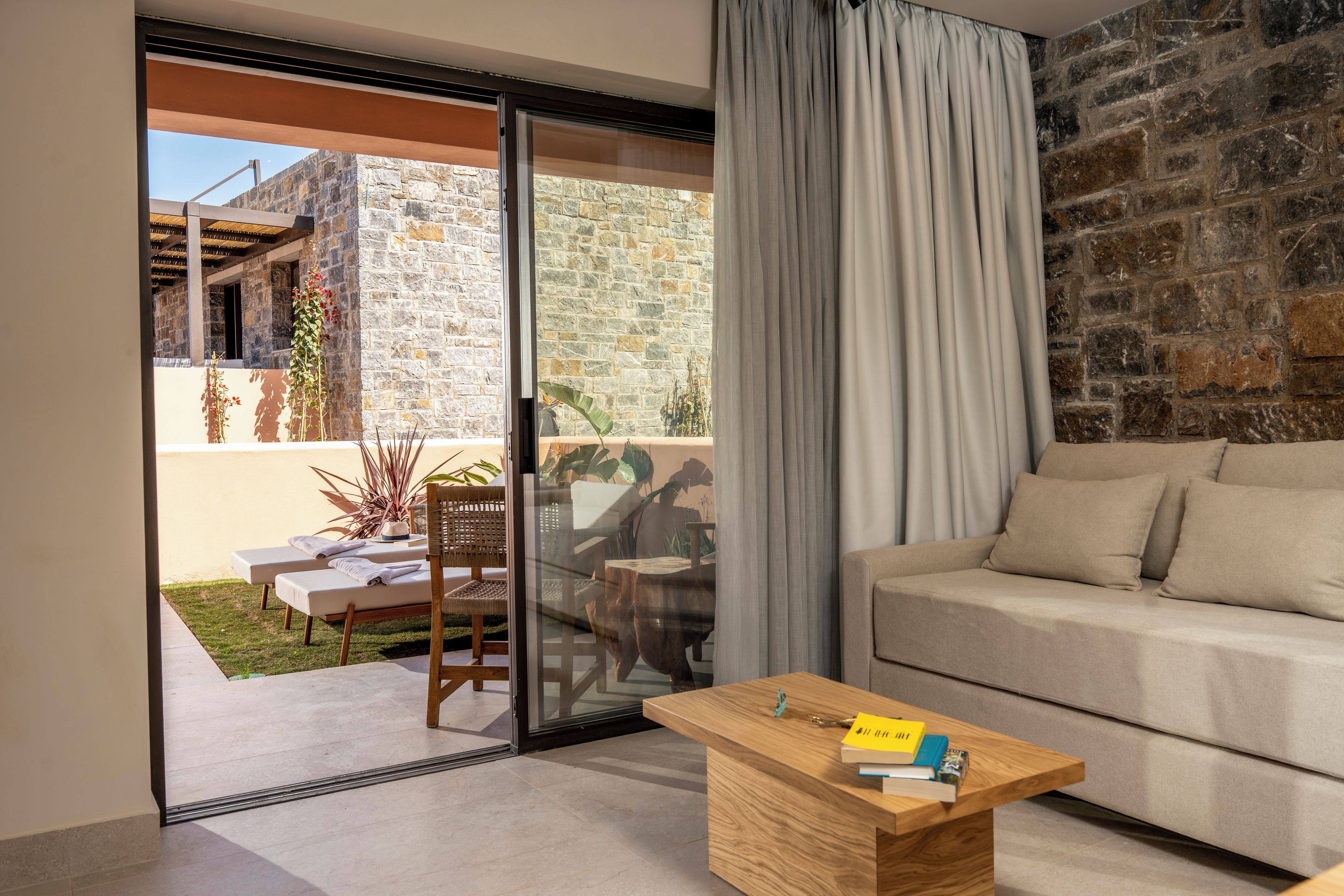 A modern living room with a sofa, a wooden coffee table, and a glass door leading to a balcony with a view of a stone building and plants.