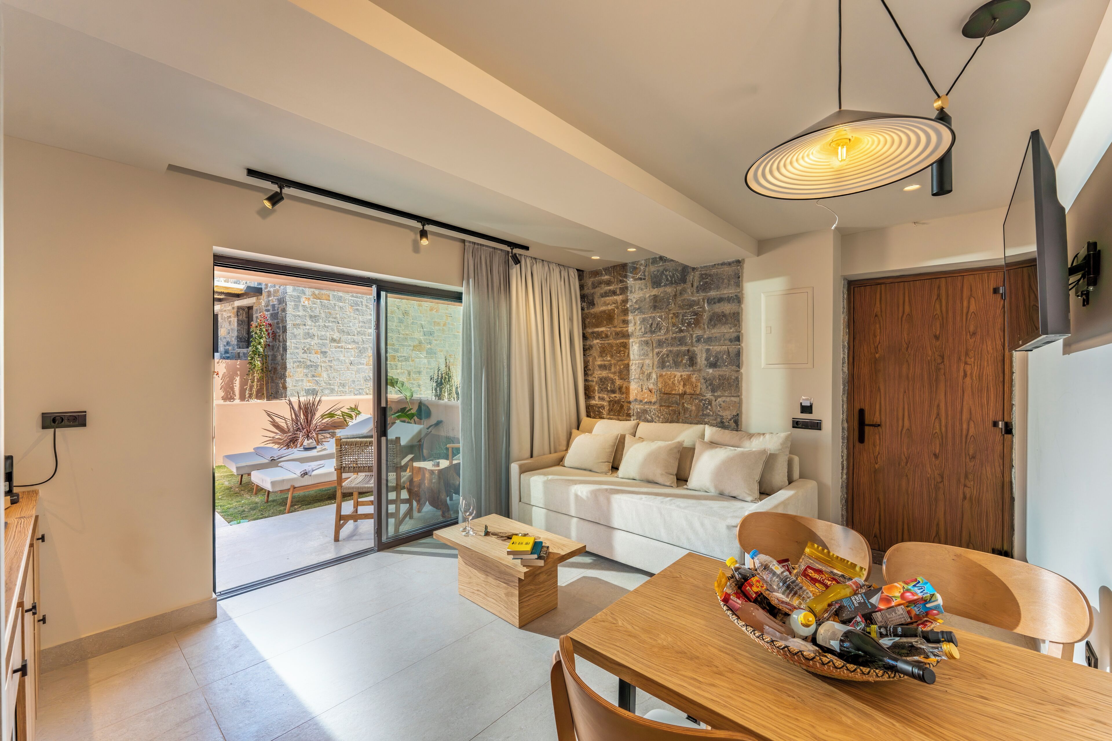 A modern living room with a sofa, a dining table with a basket of snacks, a coffee table, and a large window with curtains.
