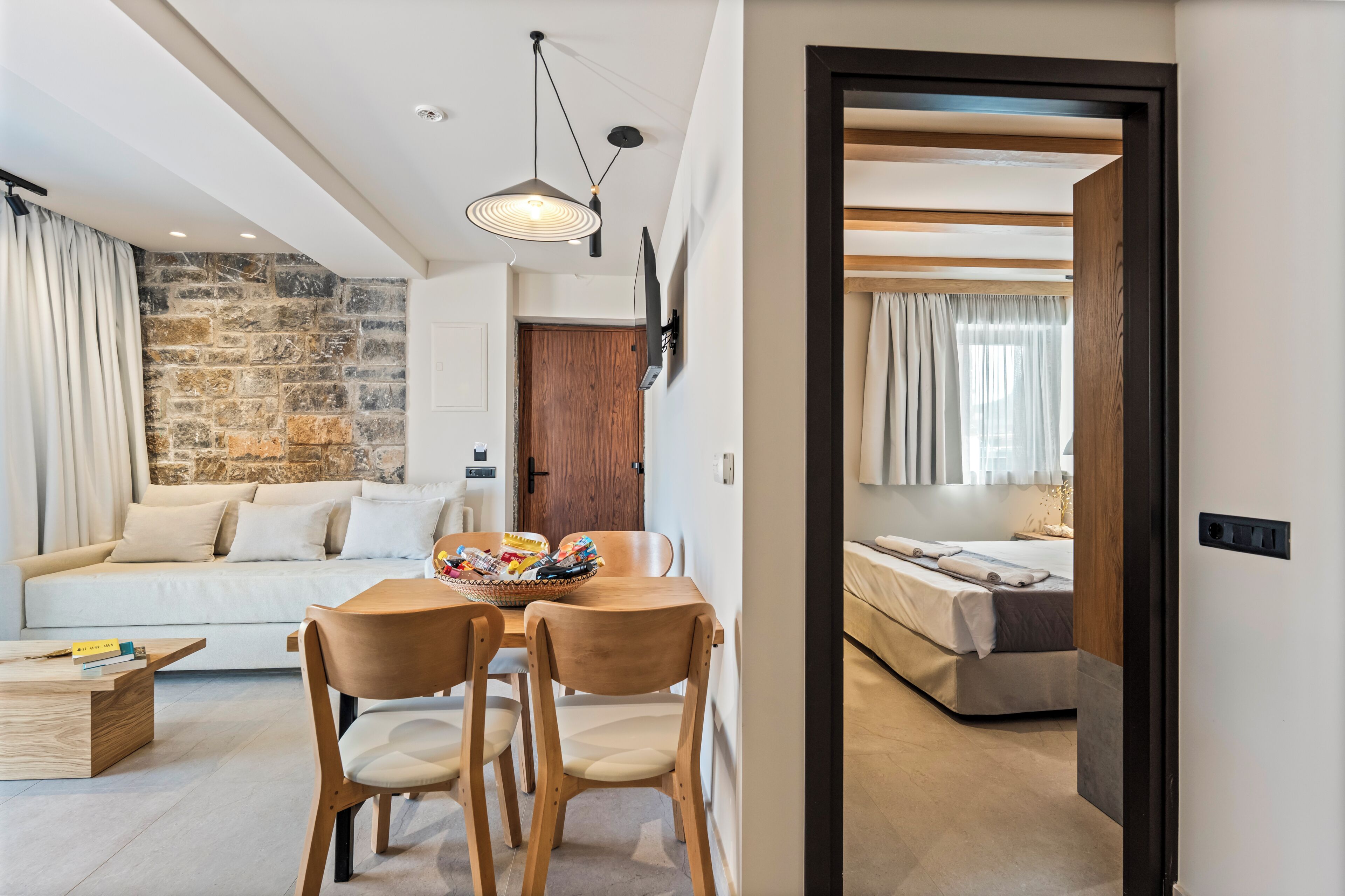 A modern living room with a stone wall, a wooden dining table with chairs, a sofa, and a bed in the adjacent room.
