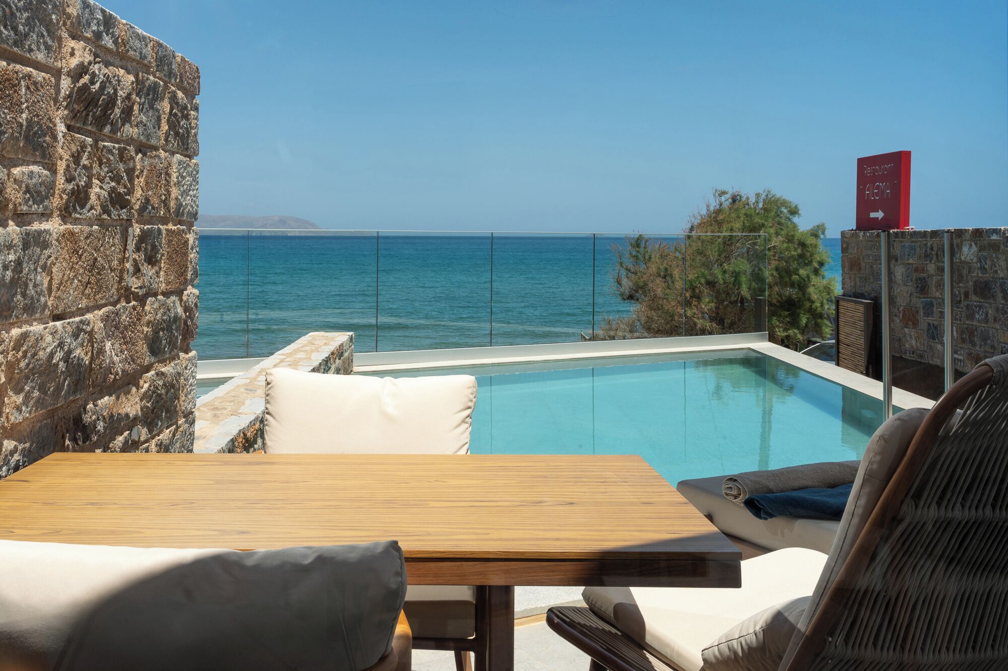 A pool with a view of the sea, a stone wall, and a table with chairs.