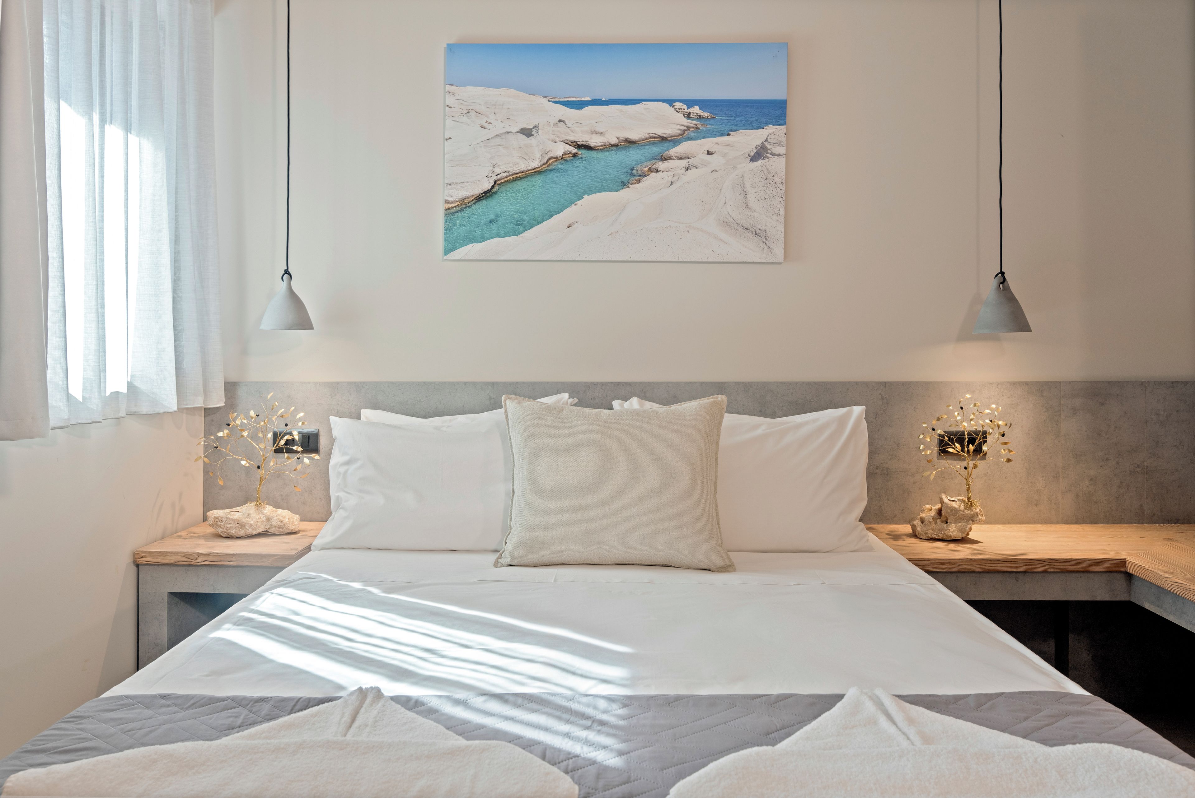A bedroom with a large bed, two bedside tables, a nightstand, a wall-mounted lamp, and a painting of a coastal landscape.