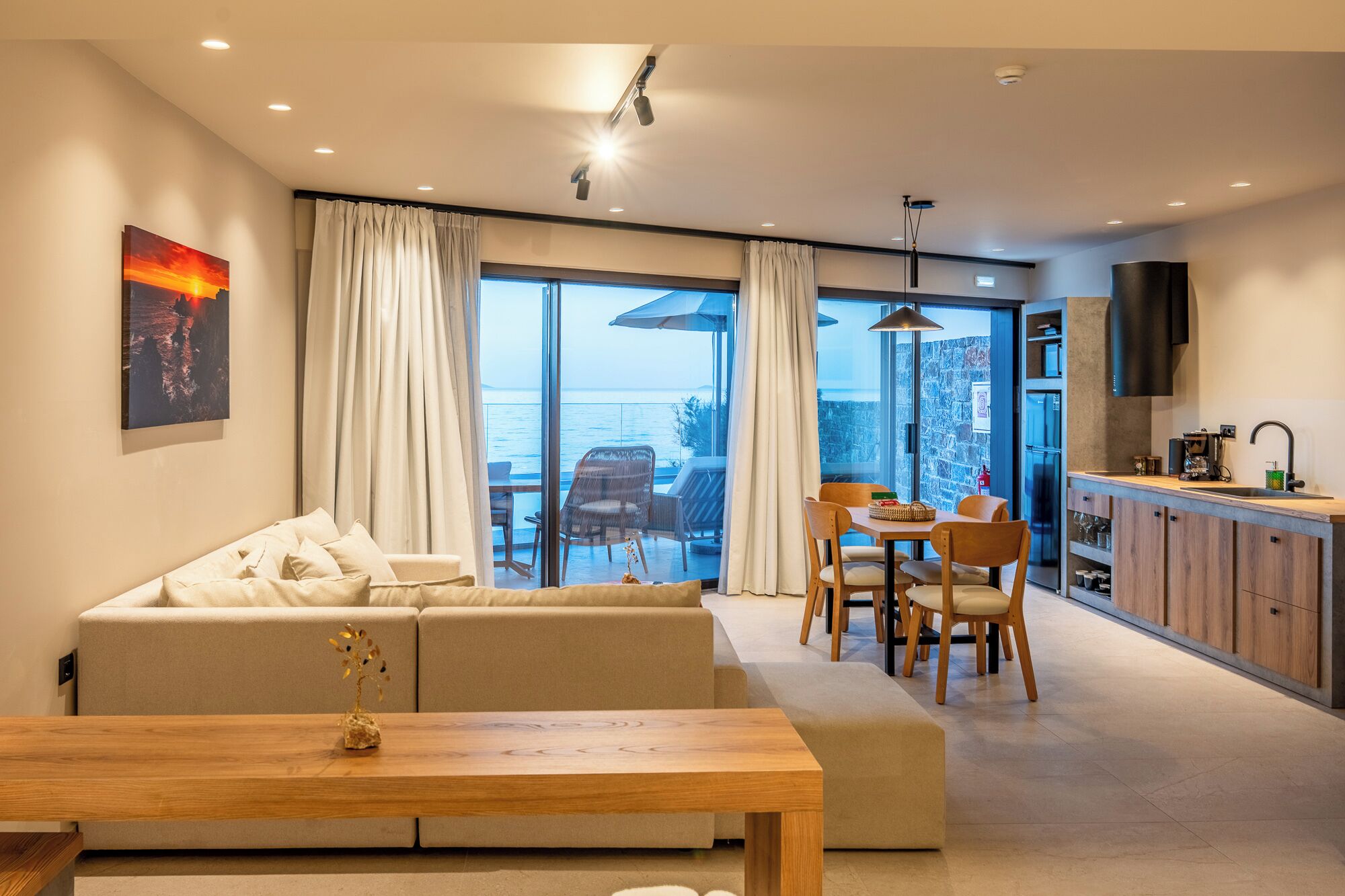 A modern living room with a sofa, dining table, and chairs, a kitchen area, and a large sliding glass door leading to a balcony with a view of the sea.