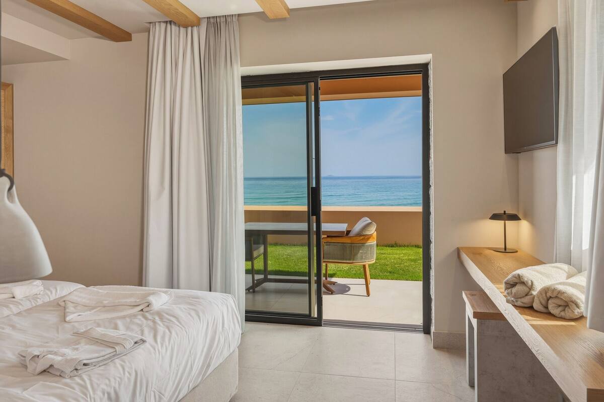 Une chambre d’hôtel avec un lit, un bureau doté d’une lampe, une chaise et une vue sur la mer, visible à travers une porte vitrée.