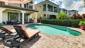 Un espace piscine avec des chaises longues, un patio couvert et une maison à deux étages dotée de balcons.