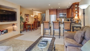 A spacious living room with a flat-screen TV, a comfortable sofa, and a coffee table. A kitchen with wooden cabinets and a microwave is visible in the background. - 1 Mi to Vail Ski Resort: Condo w/ Resort Perks! (Vail)