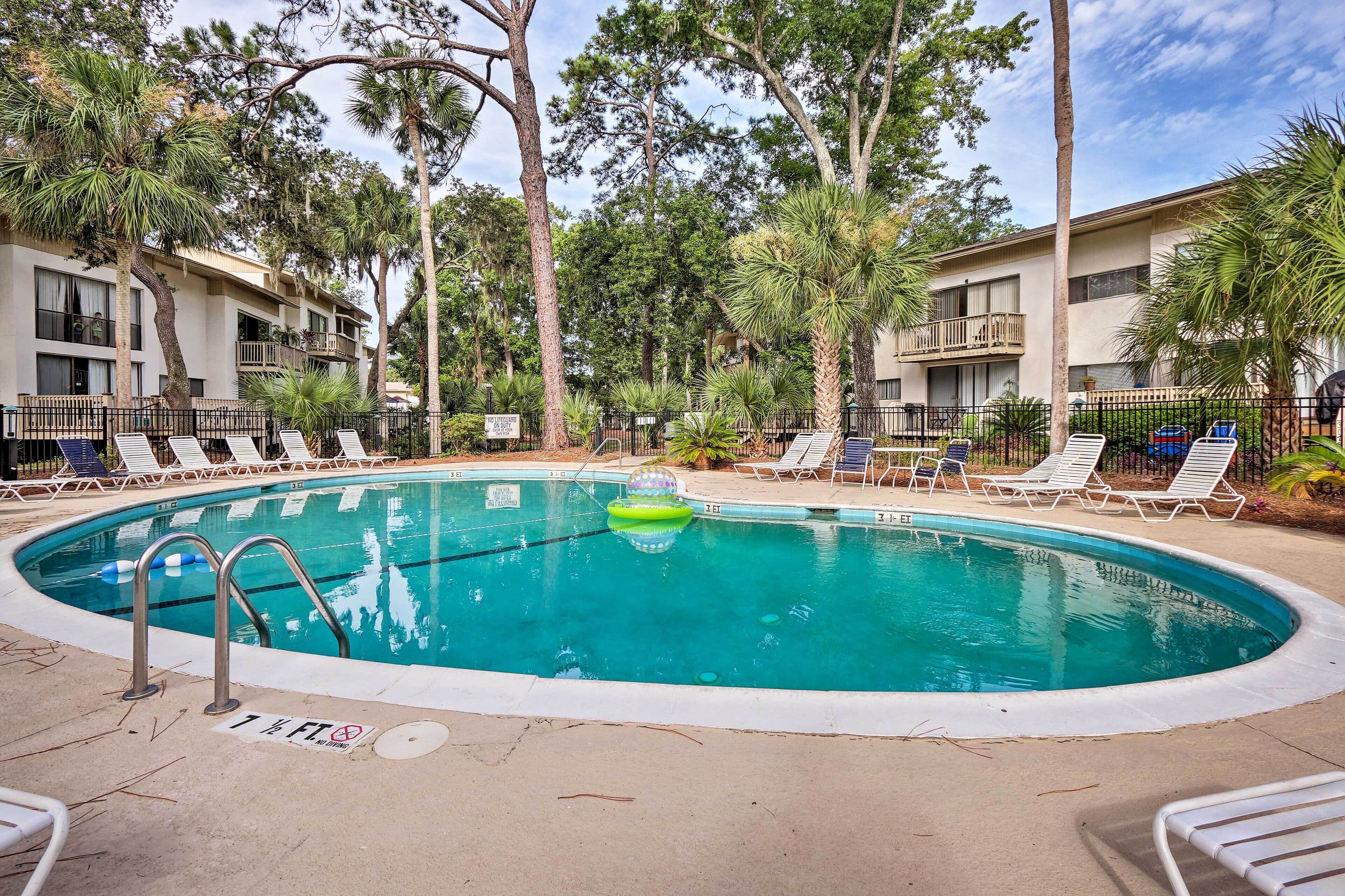 A swimming pool with lounge chairs, surrounded by palm trees and a multi-story building.