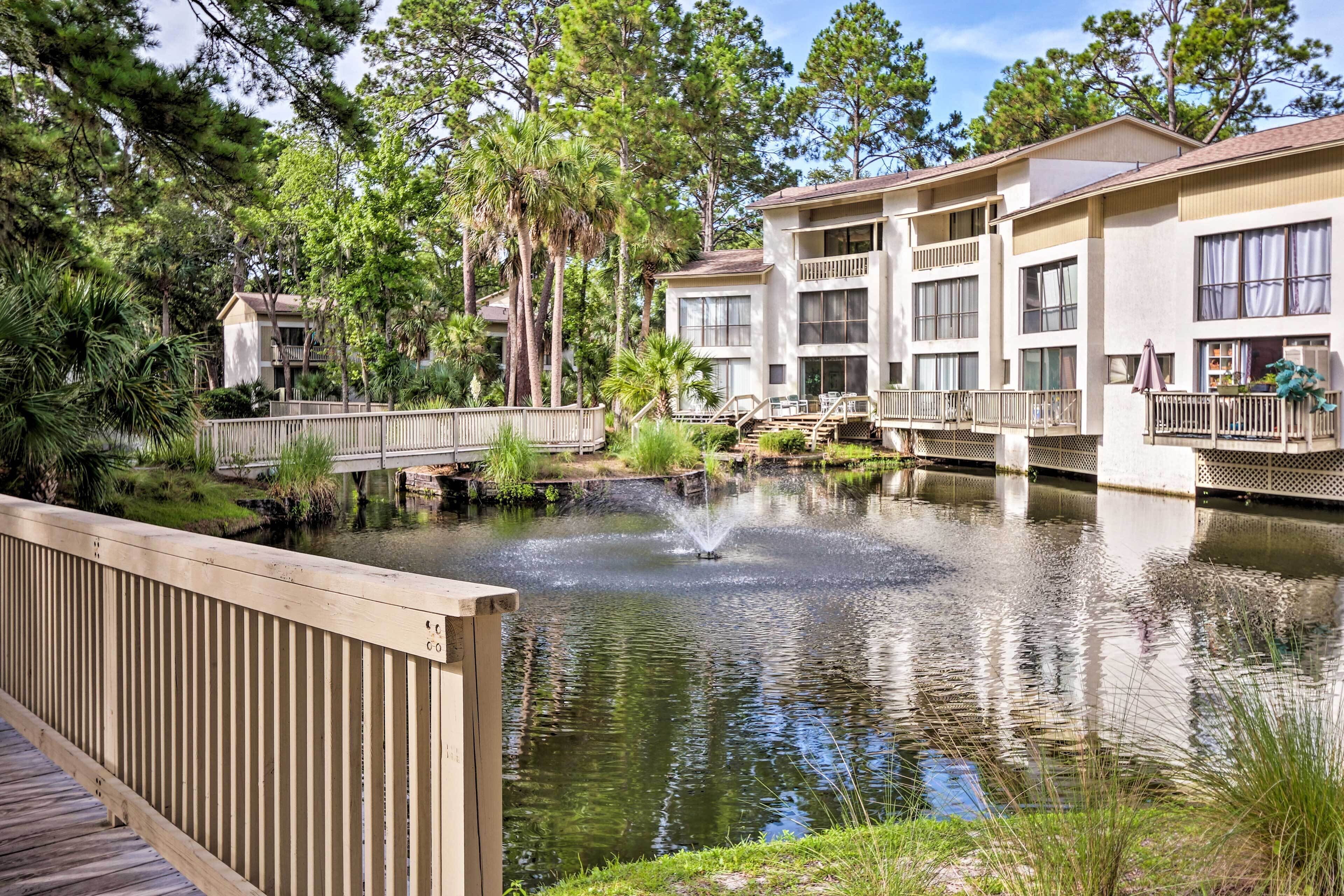 A tranquil lakeside setting with a wooden deck, a small bridge, and a building with balconies and large windows.