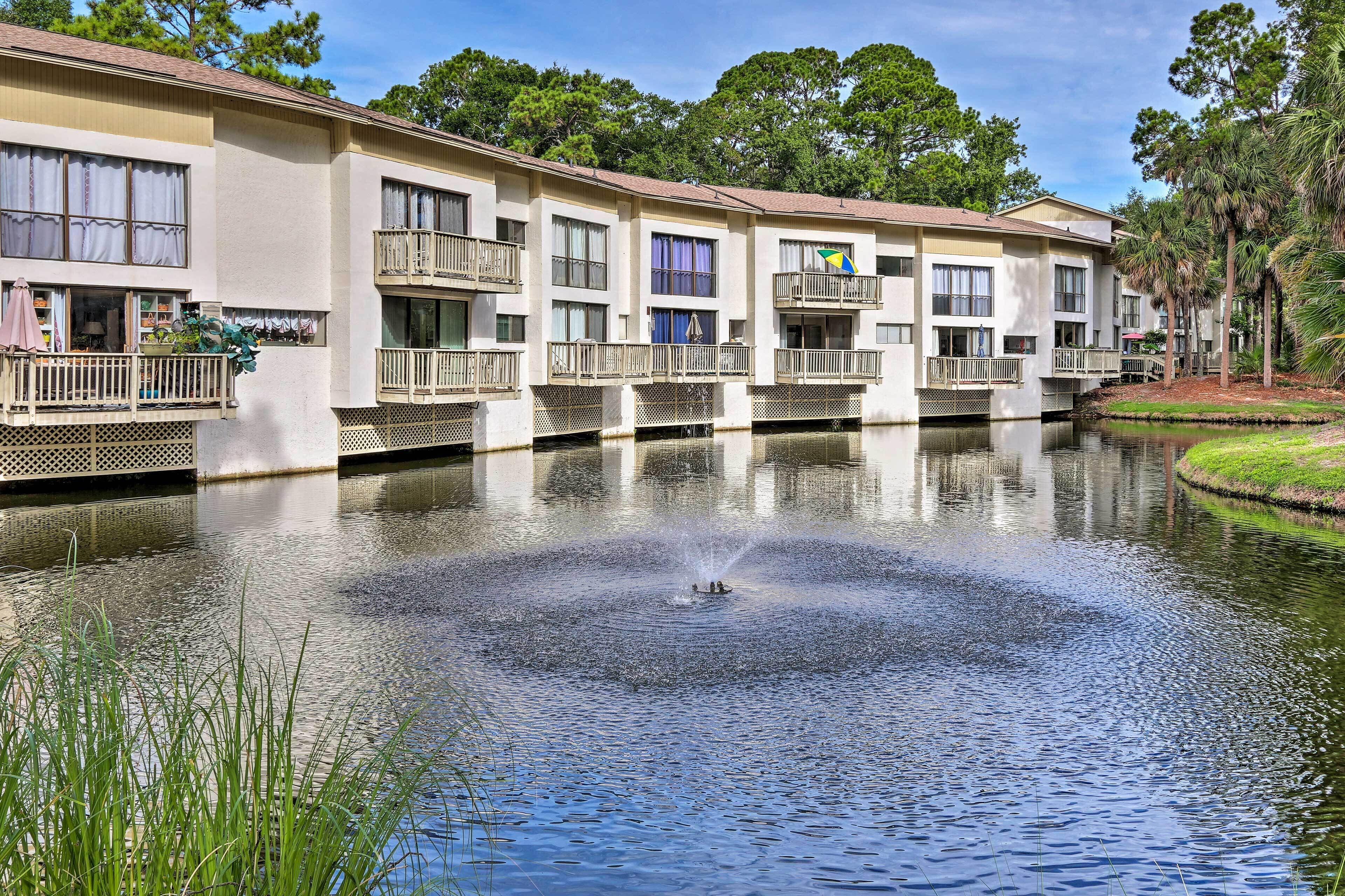 A multi-story hotel with balconies, a pond with a fountain, and surrounding greenery.