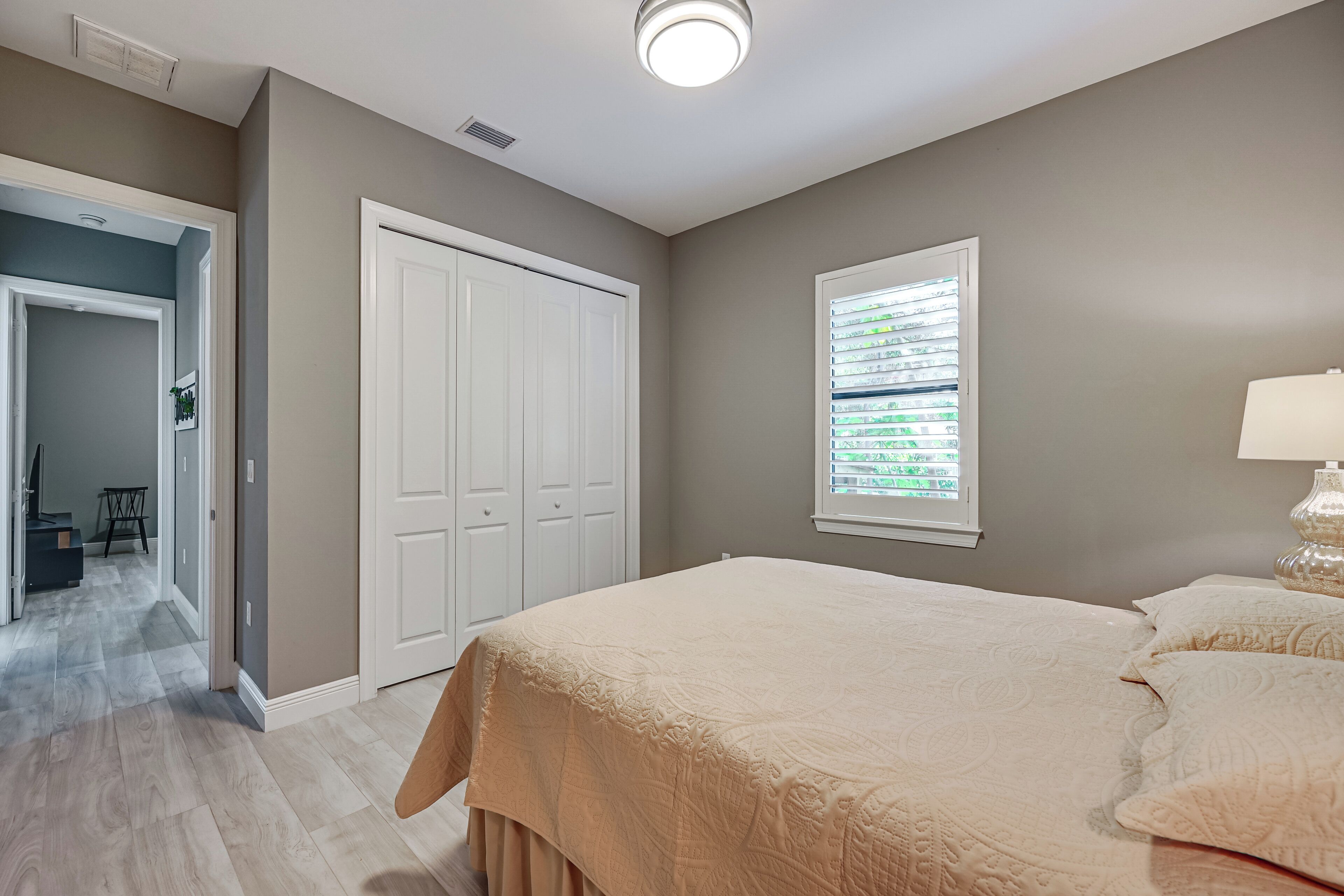 A bedroom with a large bed, a window with blinds, a white door, and a hallway leading to another room.