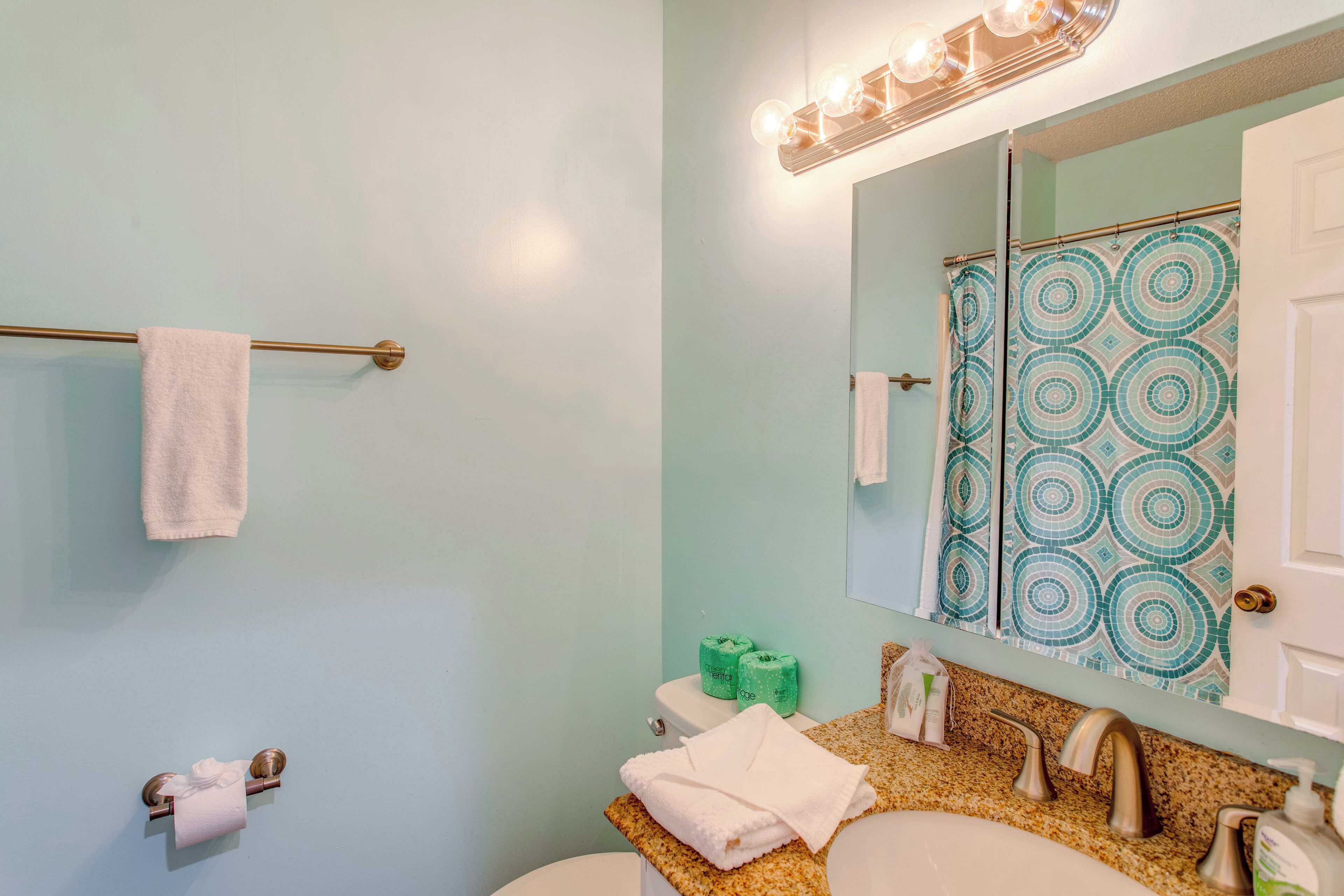 A bathroom with a light blue wall, a white sink, a mirror with a patterned shower curtain, and a towel rack.