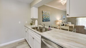 A modern kitchen with a marble countertop, stainless steel sink, and white cabinetry. There is a dining area with a table and chairs, and a living room with a couch and a chandelier. - 2 Mi to Ocean! Poolside Escape in Miramar Beach (Miramar Beach)