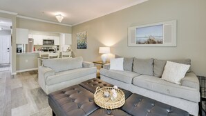 A living room with a grey sofa, a black ottoman, and a dining area with a kitchen in the background. - 2 Mi to Ocean! Poolside Escape in Miramar Beach (Miramar Beach)