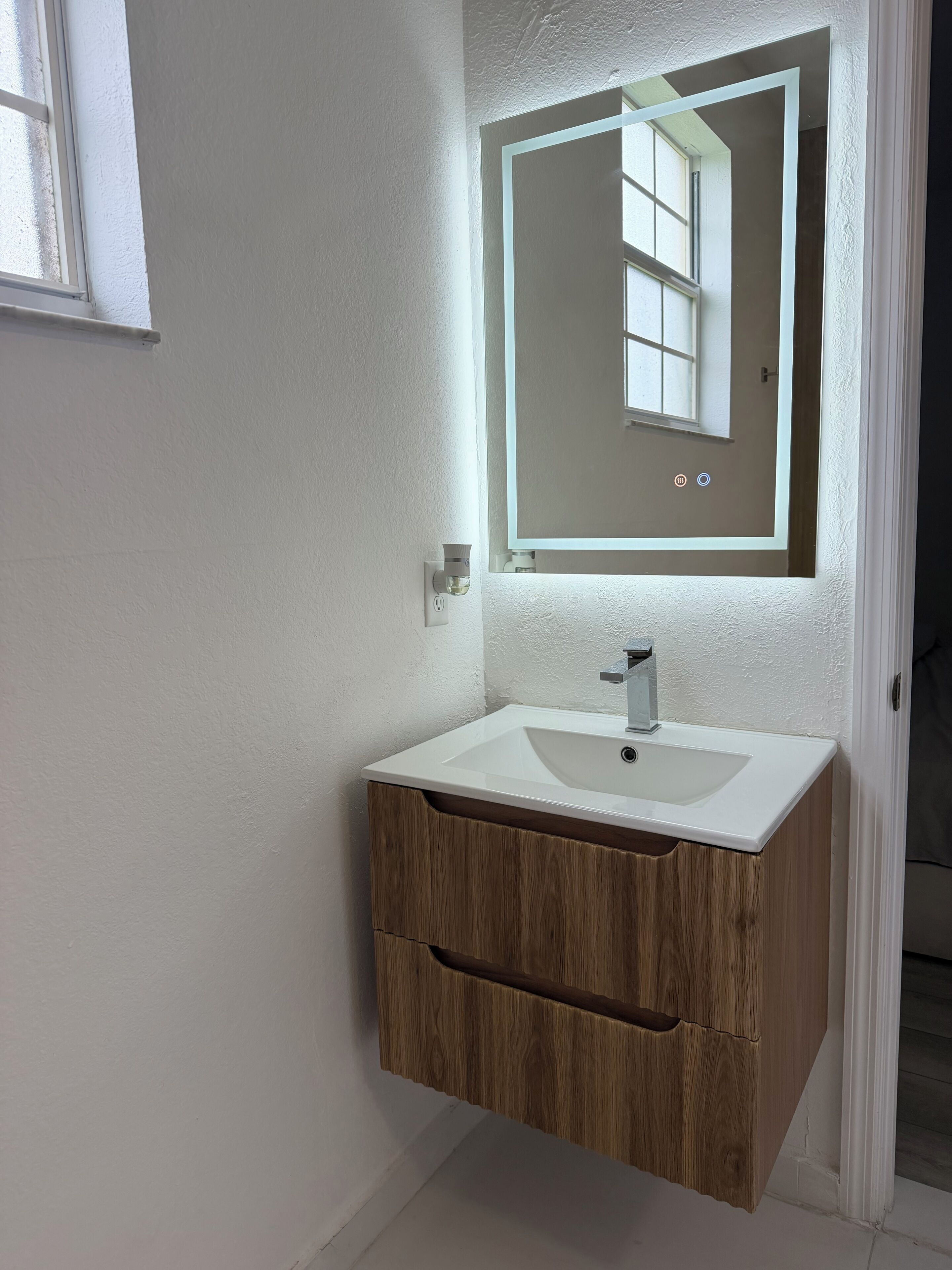 Un bagno con un lavandino bianco, mobile contenitore in legno e uno specchio illuminato.