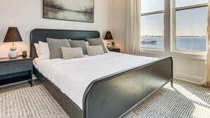 Un dormitorio moderno con una cama grande, dos mesitas de noche, una mesita auxiliar, una ventana con vista al agua y un cuadro en la pared.