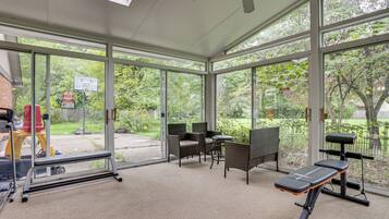 A gym with exercise equipment, wicker chairs, and large windows overlooking a garden.
