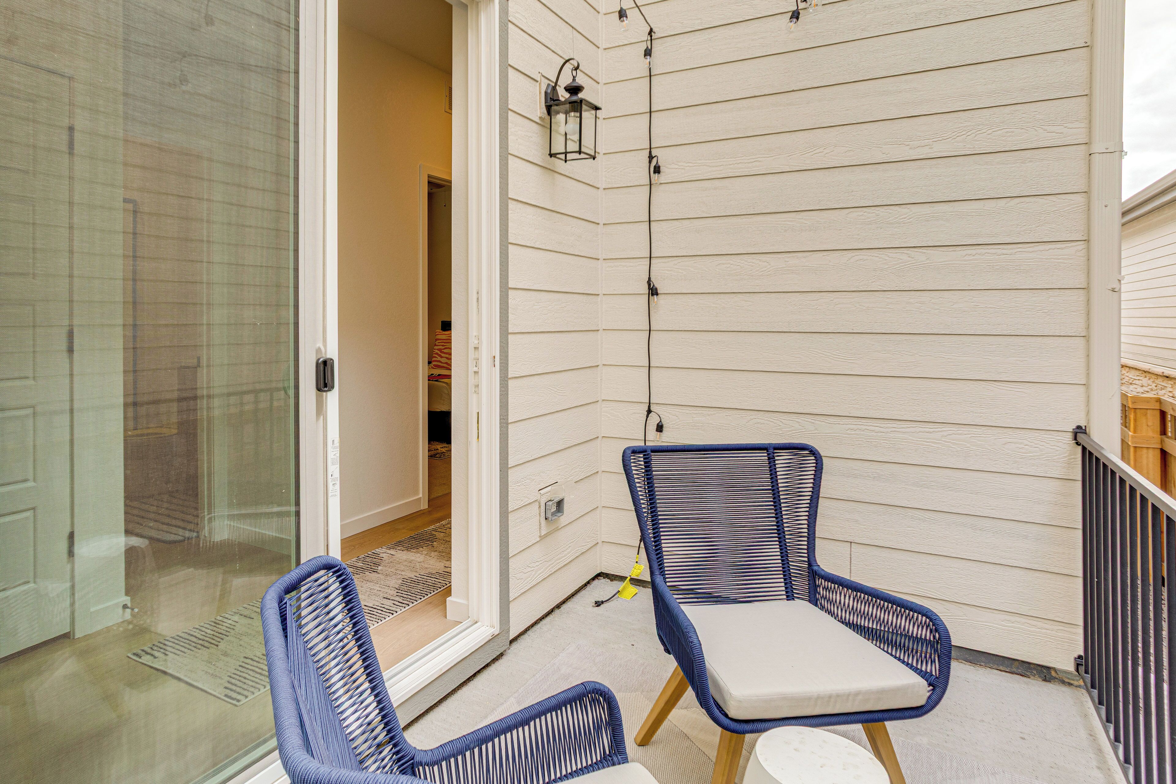 En balkon med to blå fletstole og et lille bord, en glasdør der fører ind, og gelænder af træ.