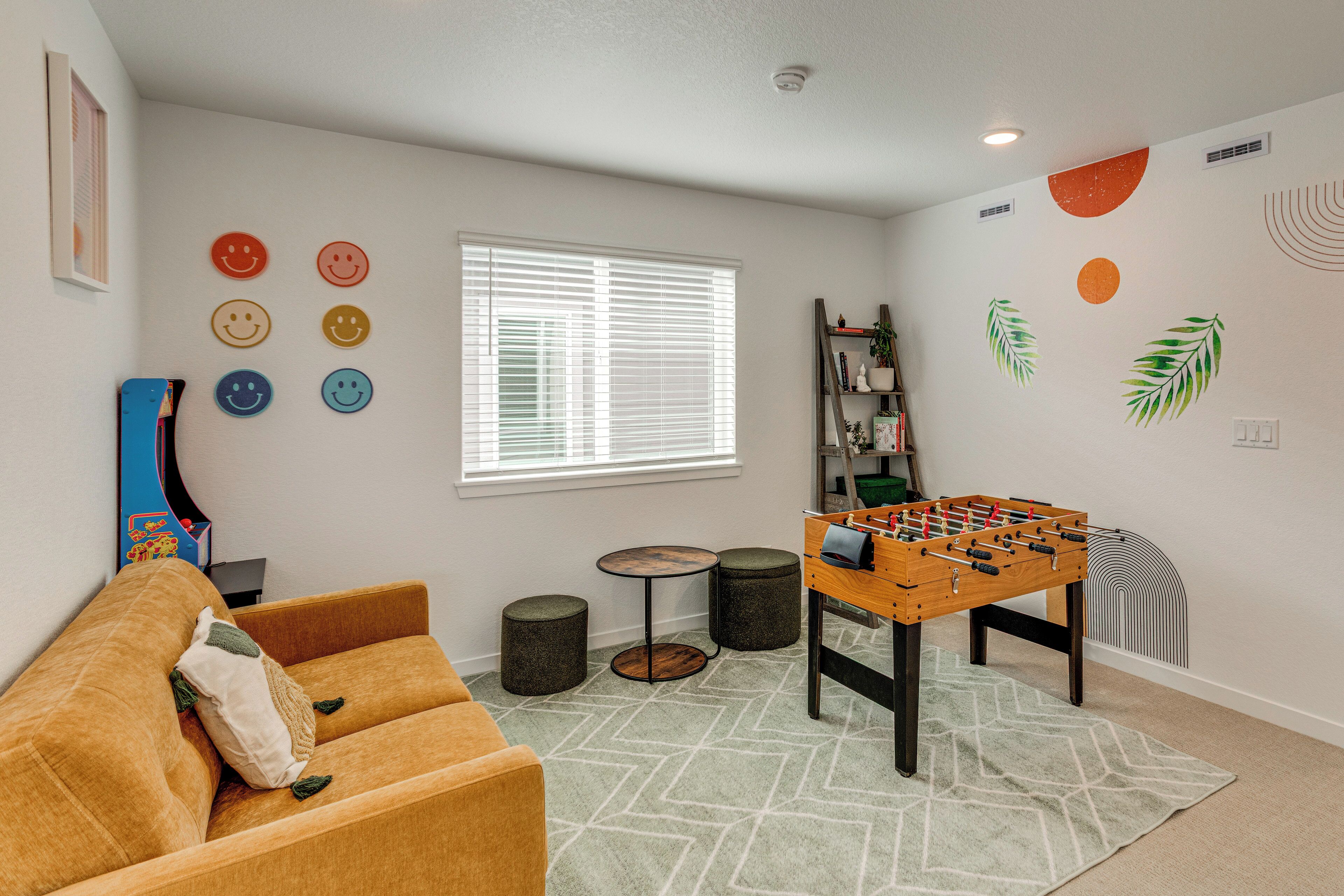 A cozy room with a foosball table, a sofa, a window with blinds, and wall decorations including smiley faces and leaf patterns.