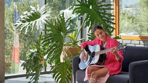 Una persona sentada en un sofá oscuro en una habitación con ventanas amplias, tocando la guitarra.