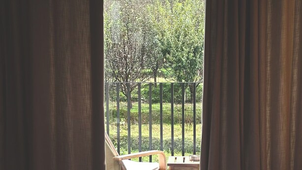 A bed with a white sheet and a black bag on it, a wooden chair, and a view of a garden through a window with curtains.